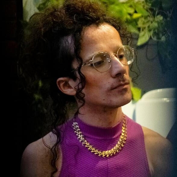 Uma pessoa com cabelo escuro cacheado preso, de óculos, colar dourado e blusa rosa-choque