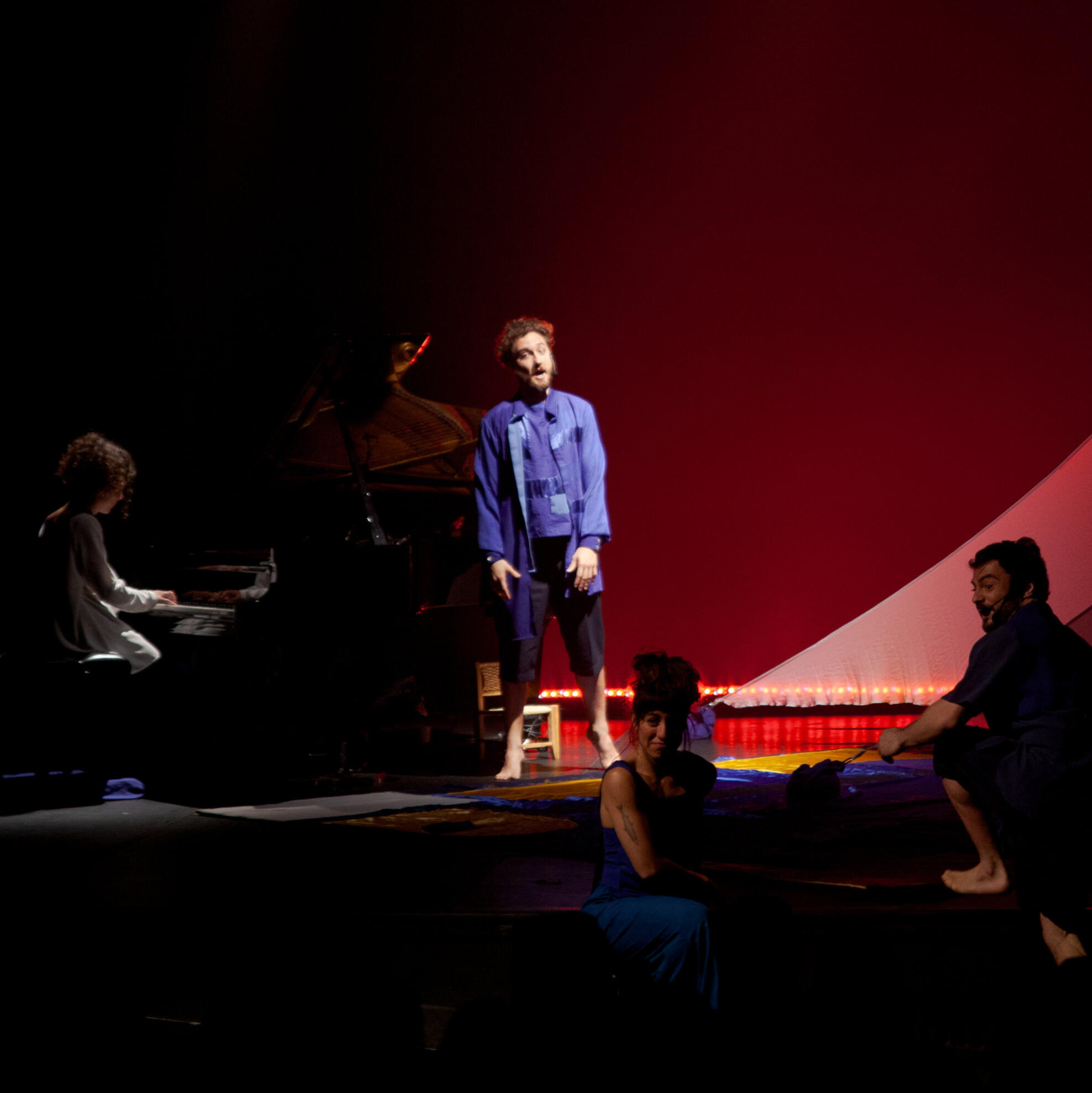 Um palco com um piano de cauda em que toca uma pianista vestida de branco, e três atores vestidos de azul